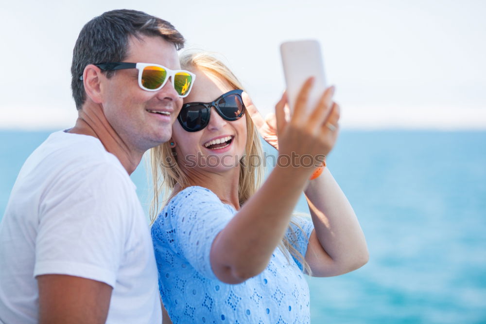 Happy young man and woman in fashionable sunglasses taking cellphone selfie on background of defocused blue sea. Vacation photos