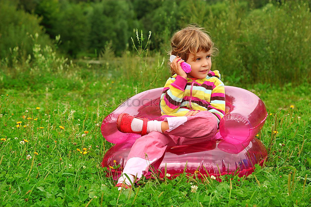 Blonde, süßes Mädchen steht mit rotem Luftballon in der Hand im Garten vor rosa Blumen in voller Blüte. Geburtstagskind draussen in der Natur freut sich über geschenkten Luftballon zu ihrem Fest.