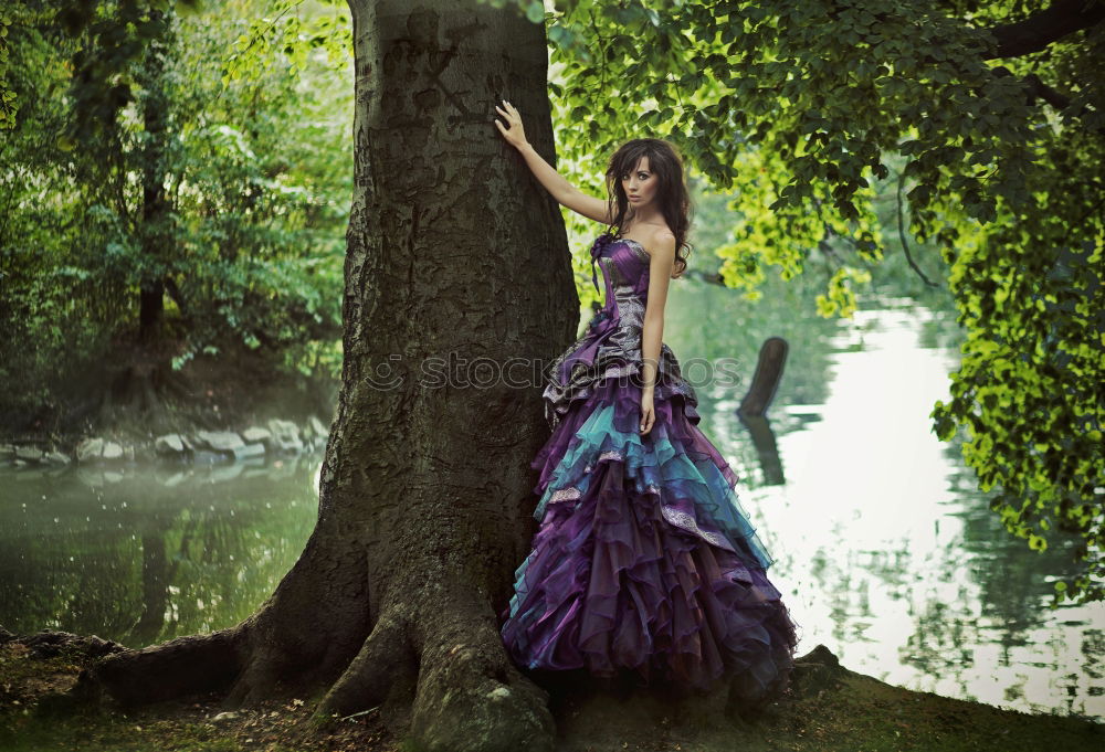 Young tall woman sitting barefoot by tree in forest in purple wedding dress