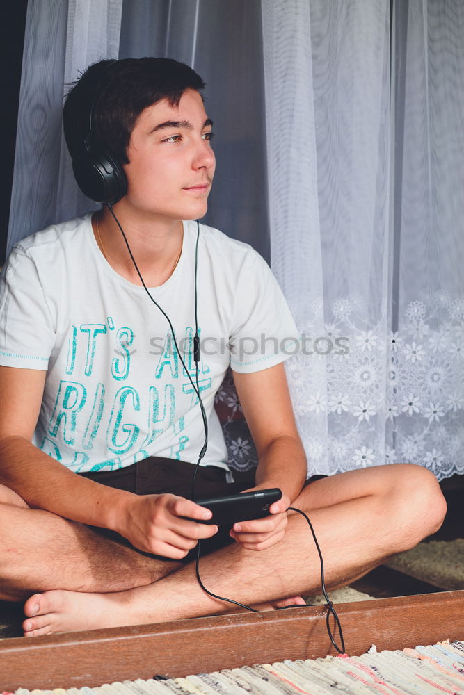 Portrait of relaxed young boy sitting at the patio door and listening to music from mobile phone