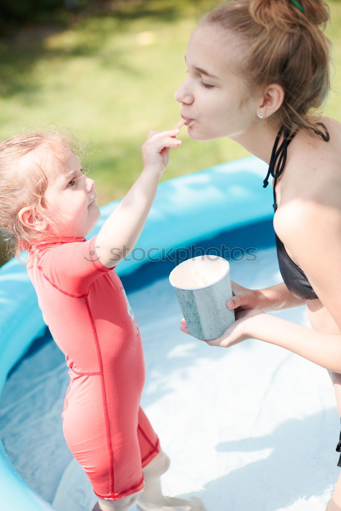 Teenage girl with her little sister spending time together in the swimming pool in a garden enjoy eating ice cream on a summer sunny day. Family quality time