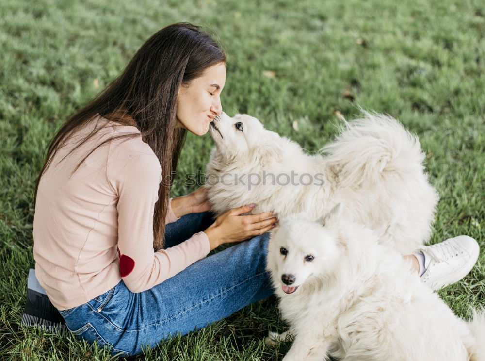 young woman with long brunette hair squats smiling on a meadow and looks at her dog