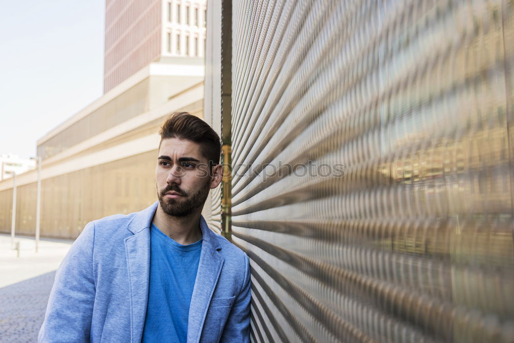 Young bearded man, model of fashion, in urban background wearing casual clothes while leaning on a wall and looking aside