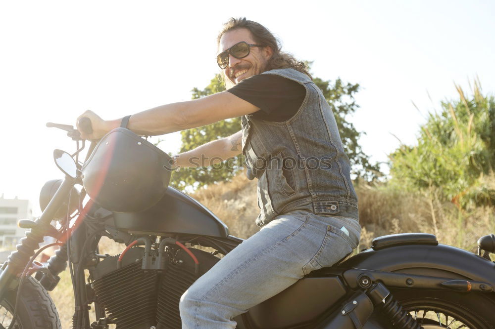 Long-haired brunette guy in sunglasses jeans and a jeans shirt posing on a black custom motorcycle