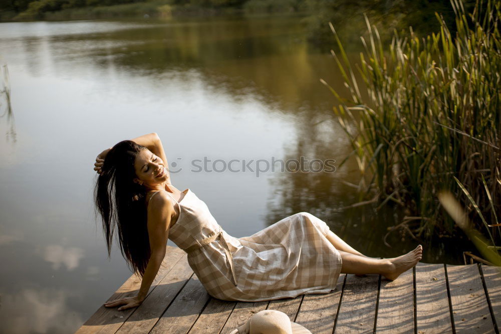 Young woman in a pink summer dress sitting in a clearing with her feet in the forest lake